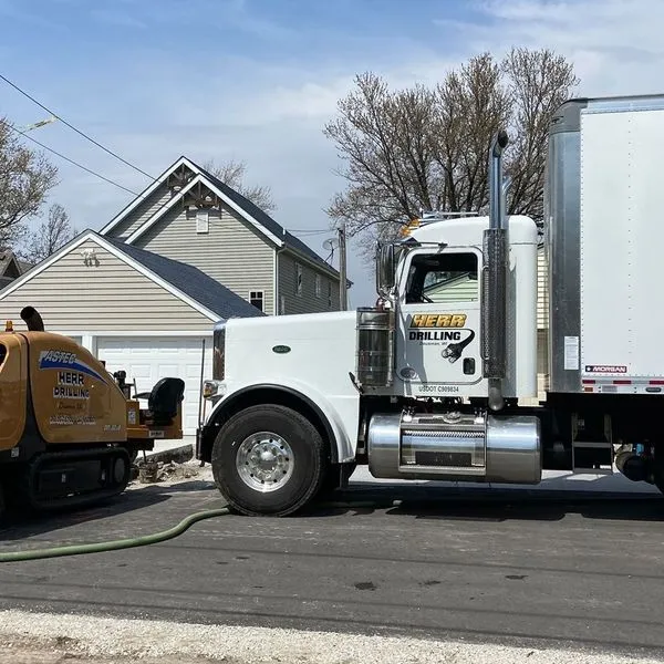 White semi-truck parked on a street next to a small construction machine, near a house.