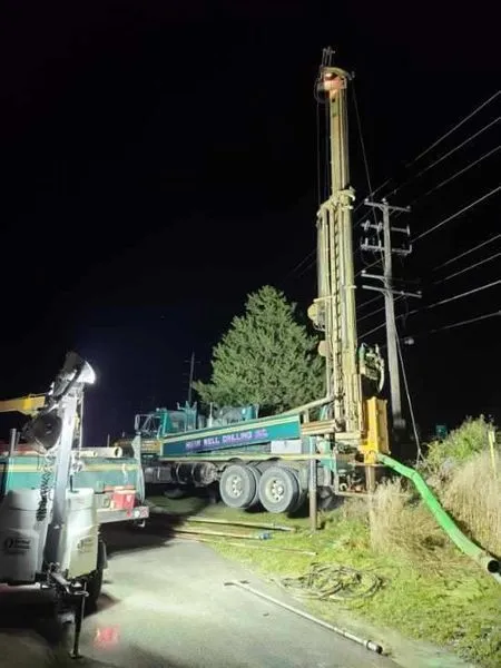 Drilling rig at night next to a power pole and greenery; green and white truck.