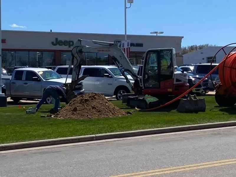 Construction worker and excavator digging on a lawn in front of a Jeep dealership, orange cable visible.
